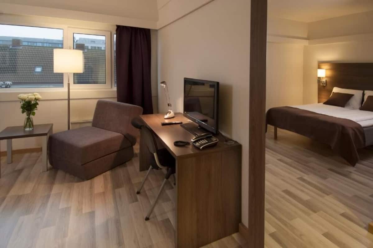 Habitación de hotel con escritorio, silla, televisión, cama y ventana con cortinas.