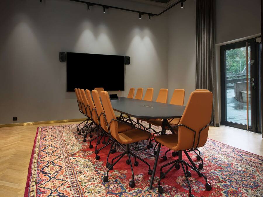 Un salón de conferencias con una gran mesa, sillas naranjas, una pantalla montada y una alfombra estampada.