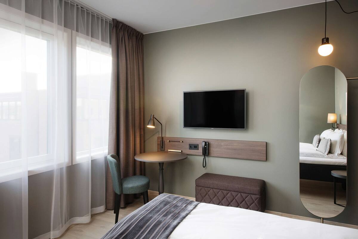 Habitación de hotel con cama, escritorio, silla, televisor, espejo y ventana con cortinas.