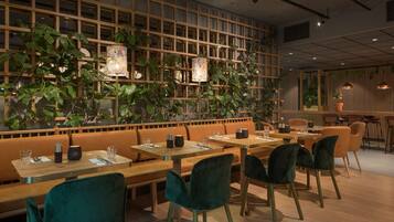 Un restaurante con mesas y bancos de madera, sillas verdes y una pared decorada con plantas y obras de arte.