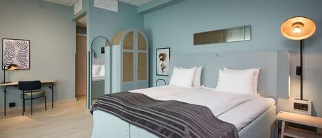 A bedroom with a bed, bedside table, lamp, desk, chair, and artwork.
