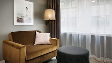 A room with a brown sofa, a black ottoman, a floor lamp, and a window with sheer curtains.