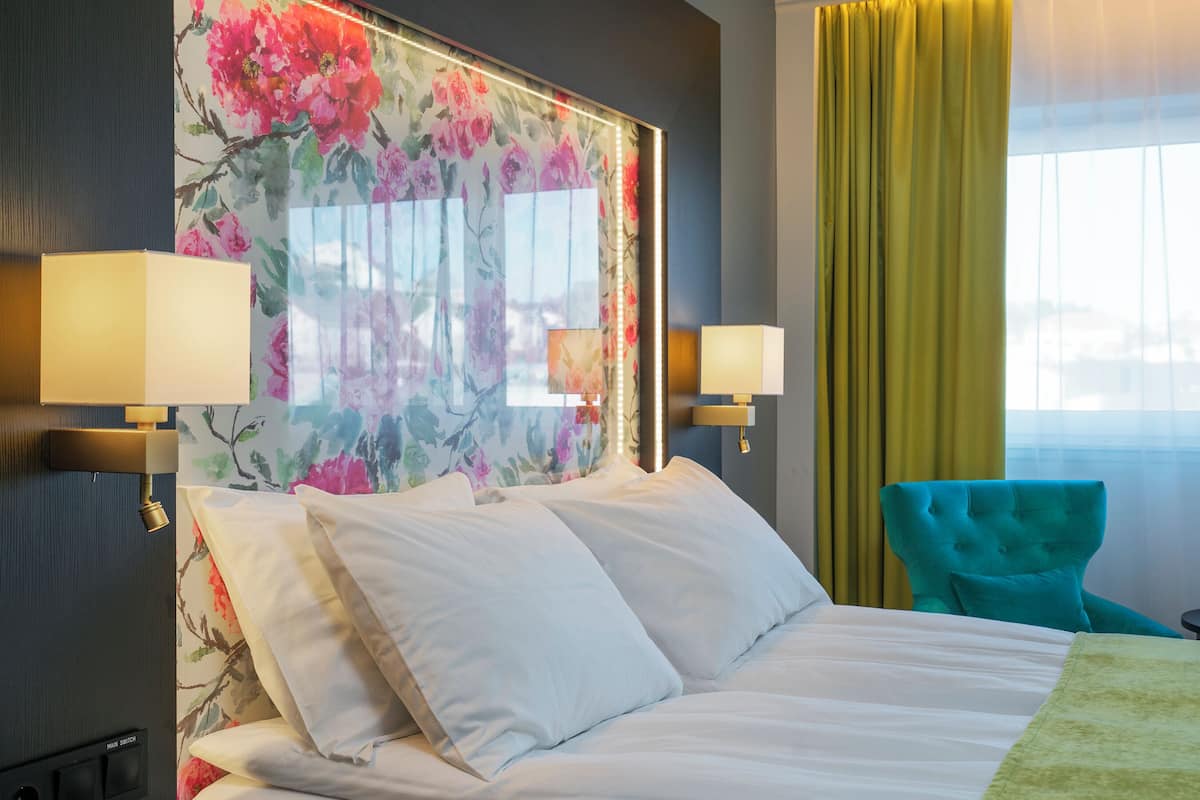 Una habitación de hotel con una cama grande, dos lámparas fijadas a la pared, un cabecero floral, un sillón azul turquesa y un ventanal con cortinas.