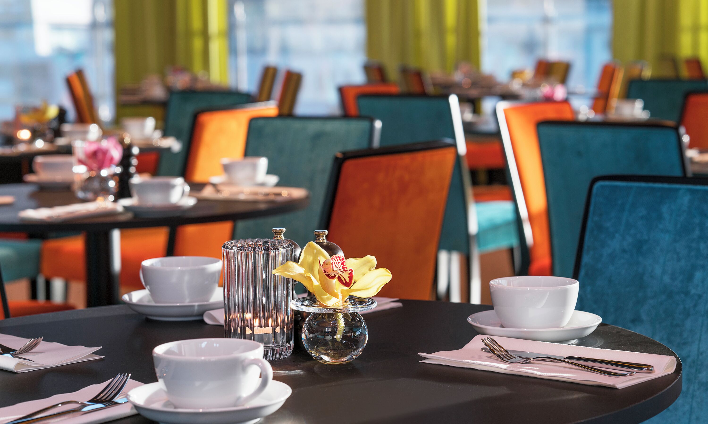 A dining area with tables set for two, featuring white cups, saucers, and a clear glass vase with a yellow flower.