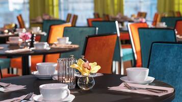 A dining area with tables set for two, featuring white cups, saucers, and a clear glass vase with a yellow flower.