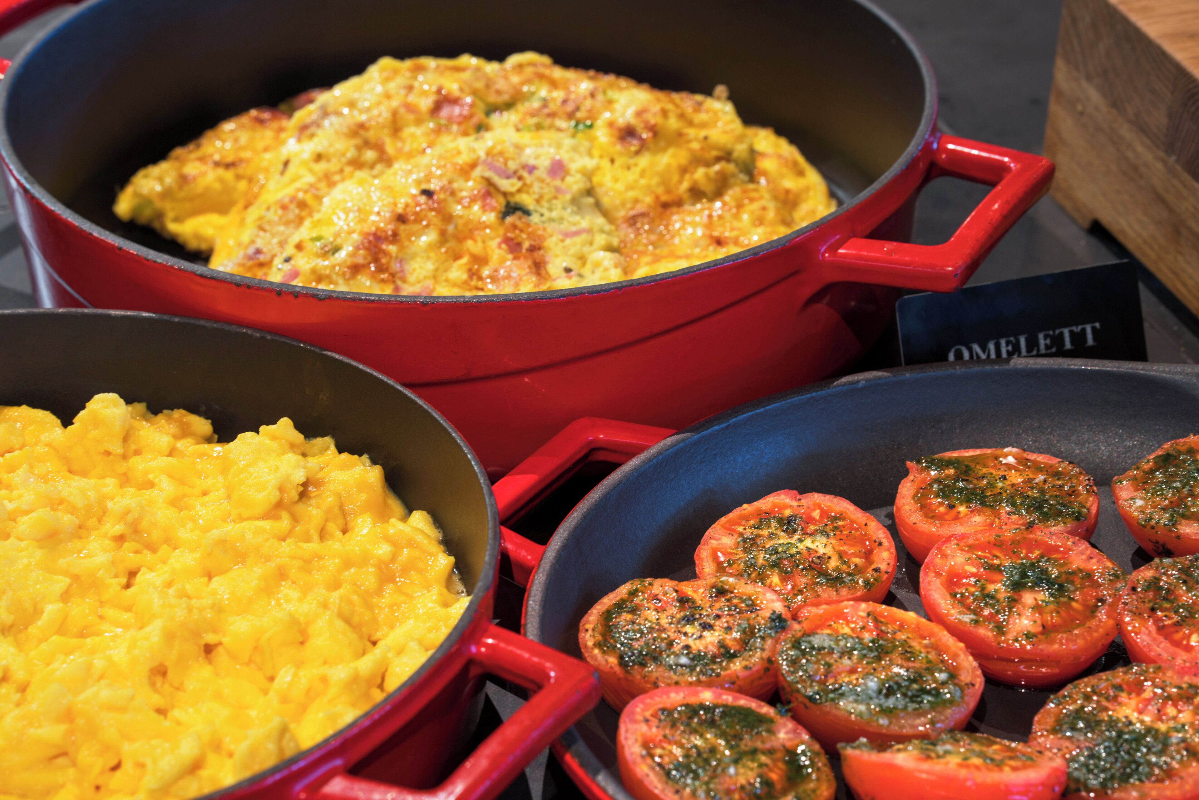 Une cuisine avec trois casseroles contenant des œufs brouillés, une frittata et des tomates au four.