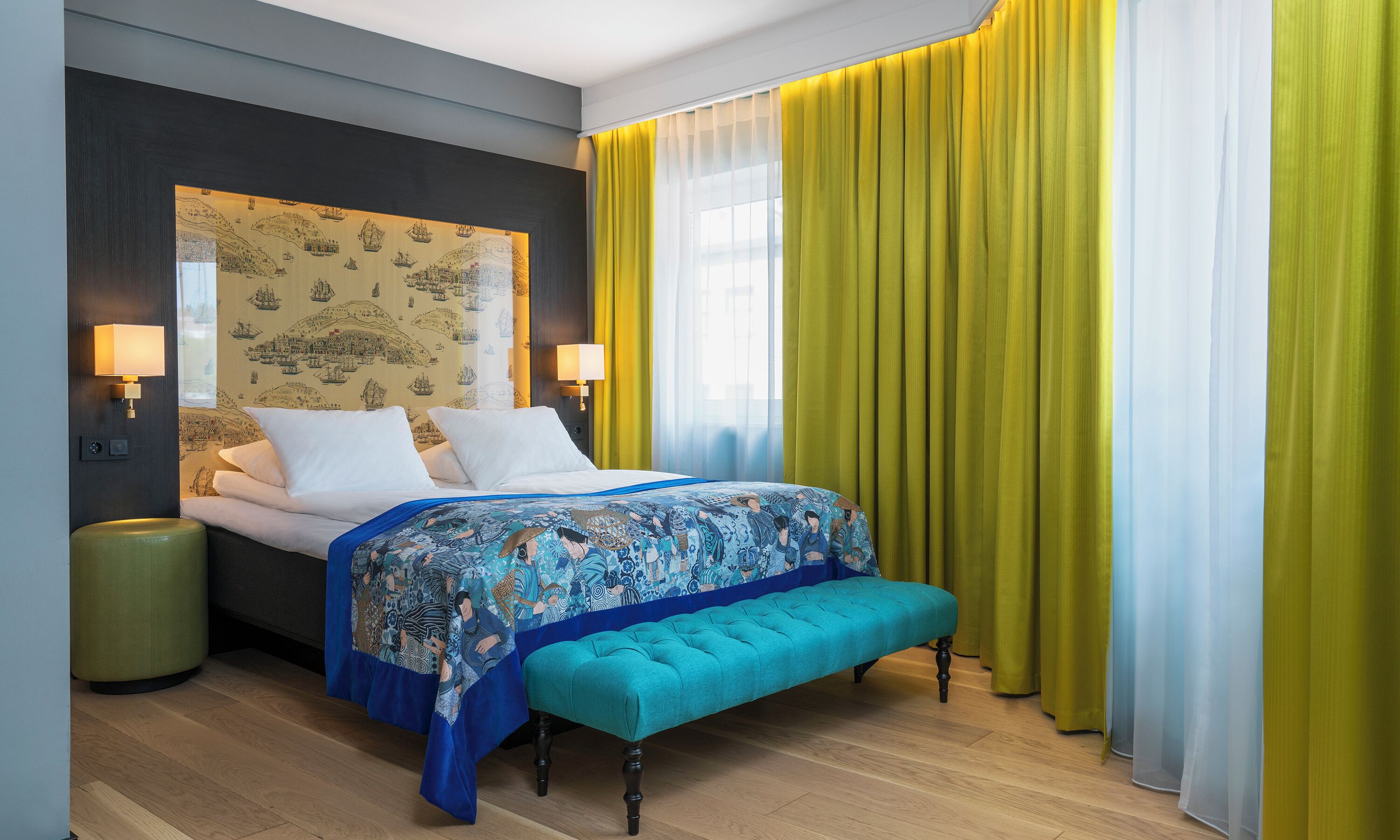 Una habitación de hotel con una cama, lámparas de noche, un banco y un ventanal con cortinas.