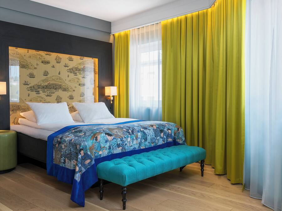 Una habitación de hotel con una cama, lámparas de noche, un banco y un ventanal con cortinas.
