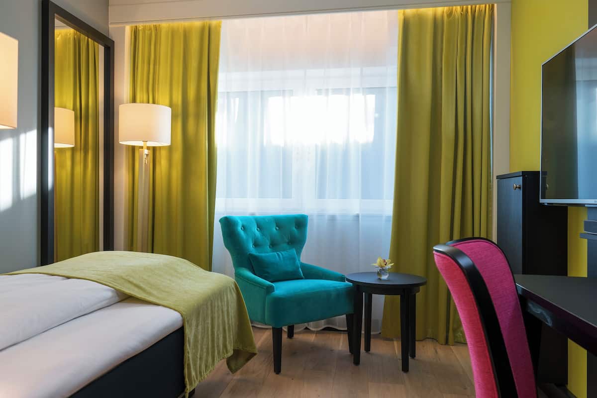 Habitación de hotel con cama, un sillón verde azulado, una mesita redonda con un florero y un televisor.