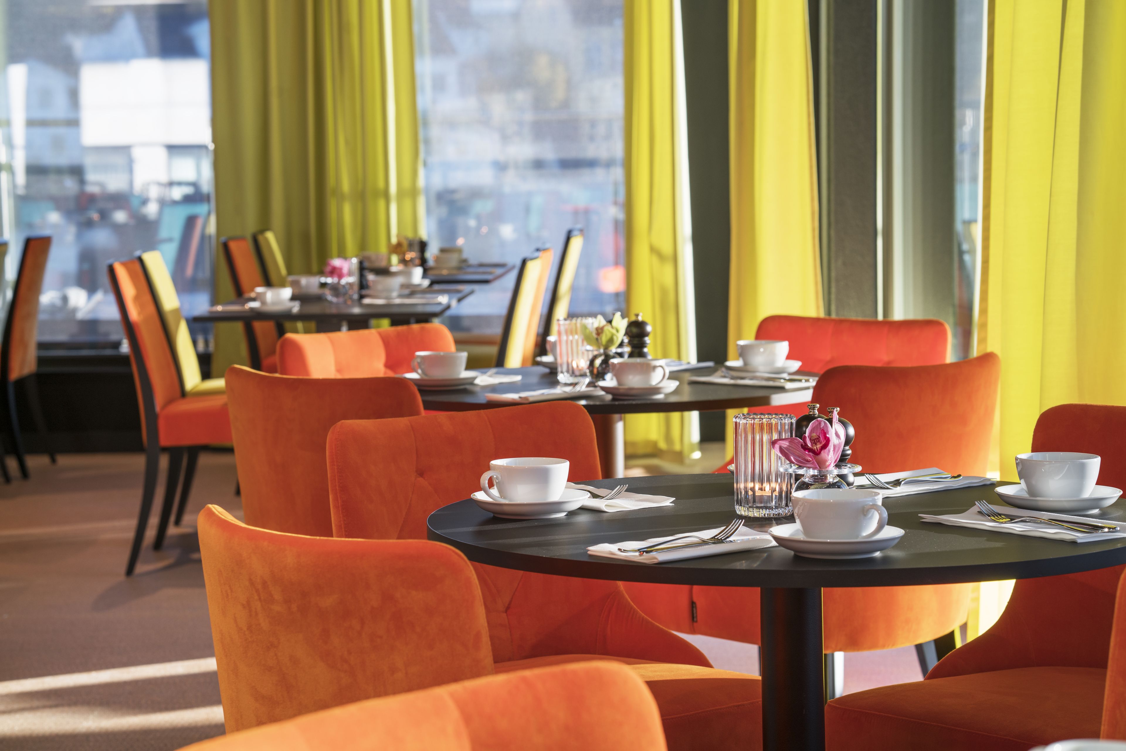 Un restaurante con sillas y mesas anaranjadas, cortinas amarillas y vistas a la ciudad a través de amplios ventanales.