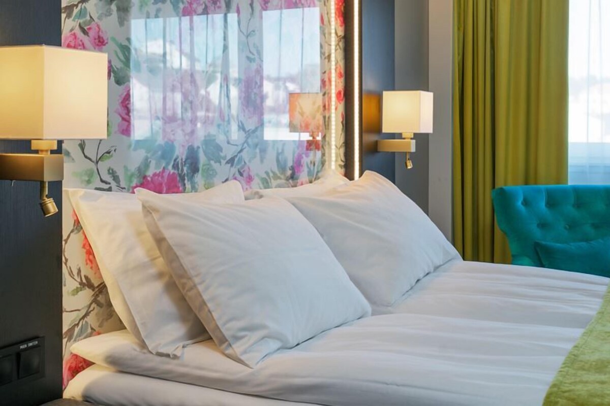 Quarto de hotel com cama, duas luminárias fixadas na parede, cabeceira com padrão floral, poltrona azul-turquesa e vista para o exterior através de uma janela com cortinas amarelas.