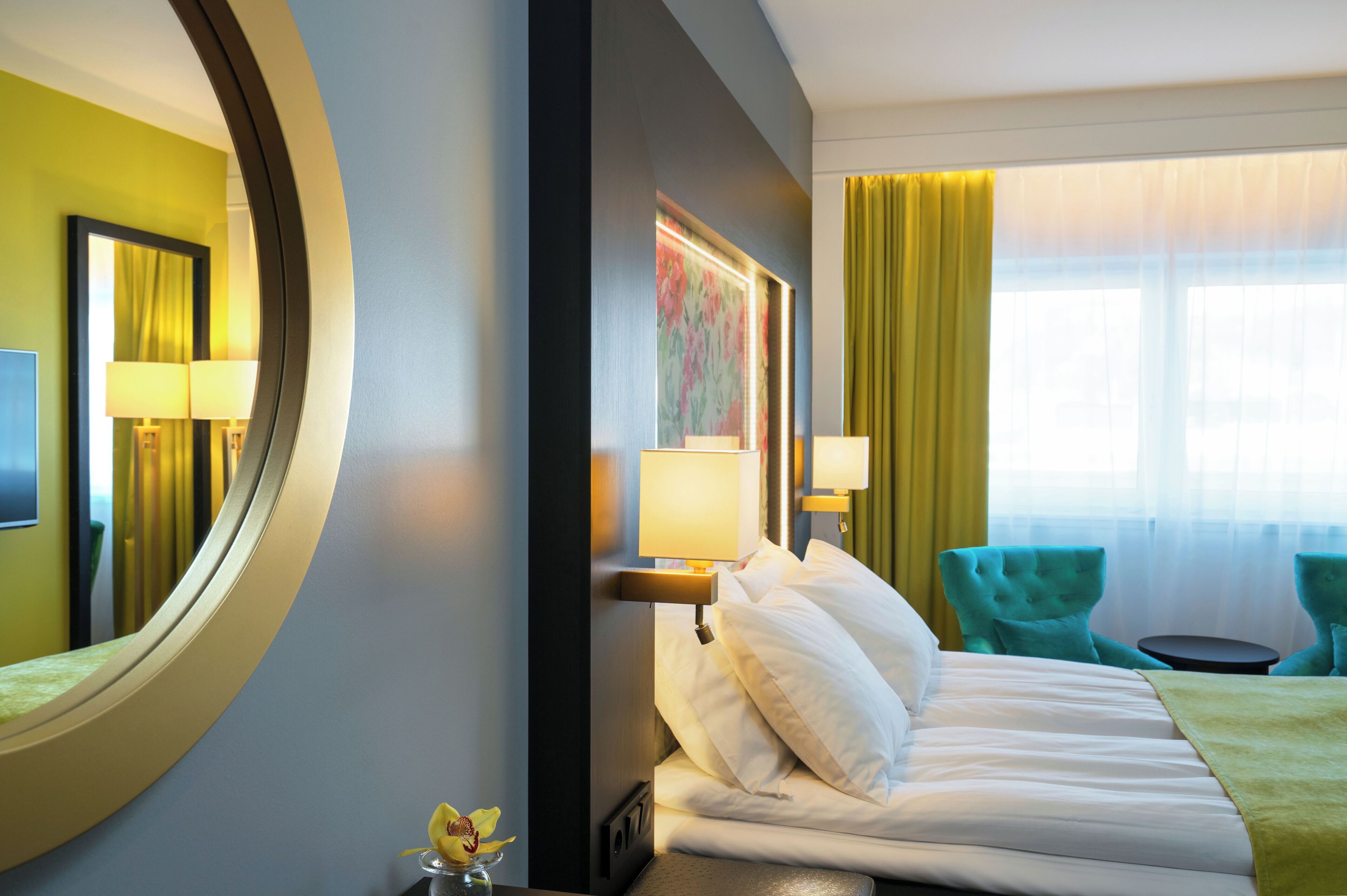 A hotel room with a bed, two chairs, a round mirror, and a window with curtains.
