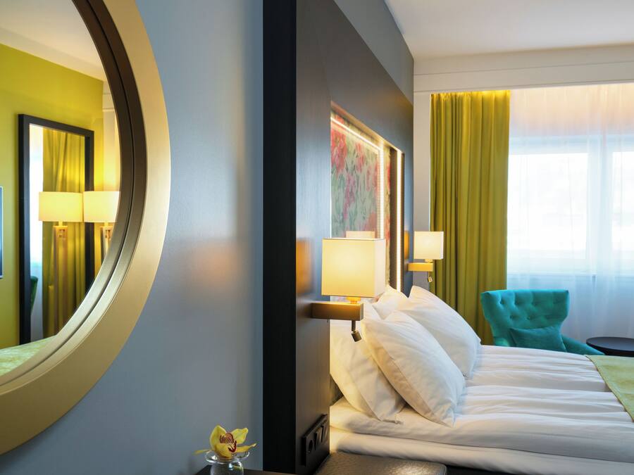 A hotel room with a bed, two chairs, a round mirror, and a window with curtains.