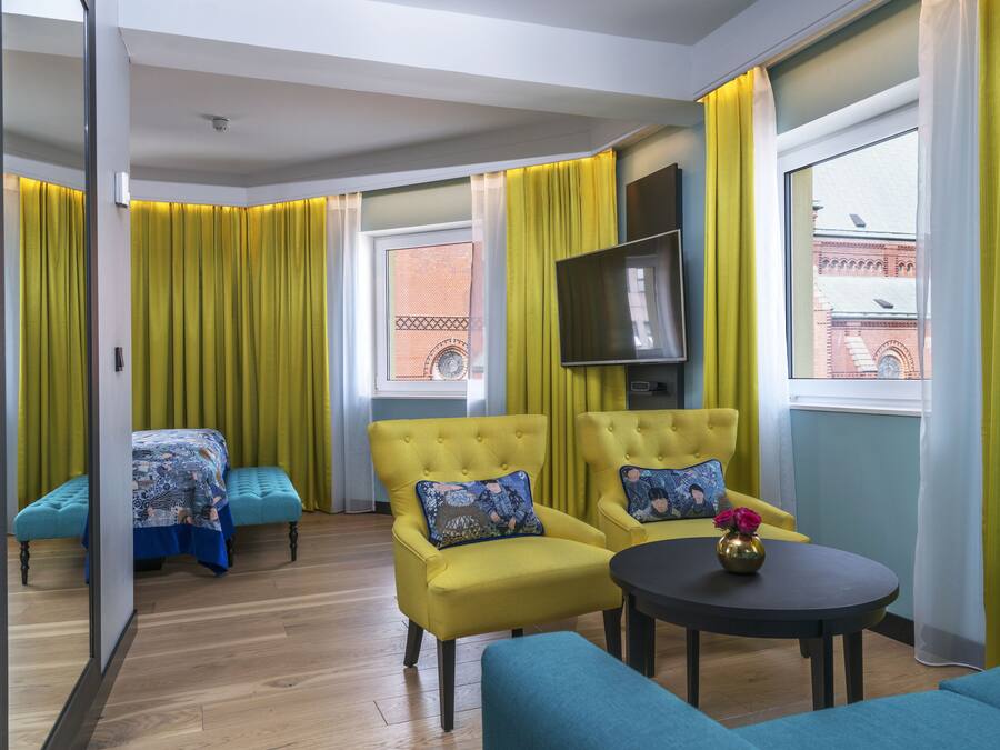 A hotel room with two yellow armchairs, a small table with a vase, a bed with a patterned bedspread, and a TV on the wall.
