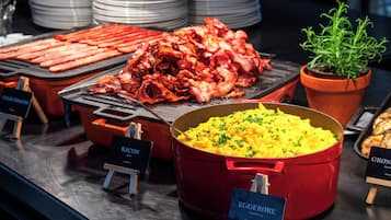 Ein Buffet mit verschiedenen Speisen, darunter Speck, Rührei und ein roter Topf mit Kartoffelpüree.