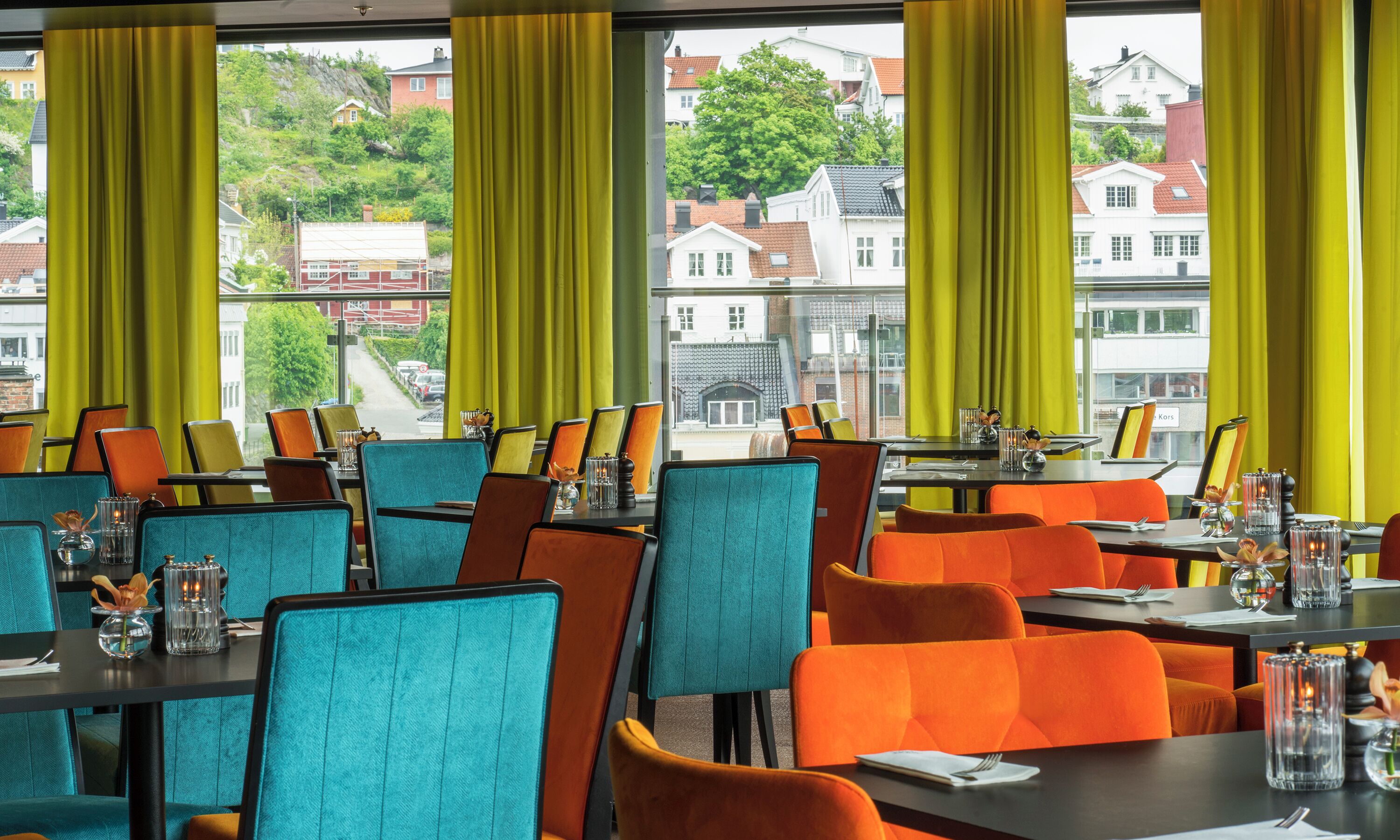 Un restaurante con mesas y sillas dispuestas para comer, amplias ventanas con cortinas y vistas a edificios y árboles.