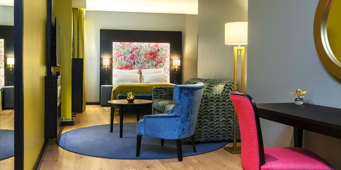 A hotel room with a bed, a blue armchair, a red armchair, a small table, and a lamp.