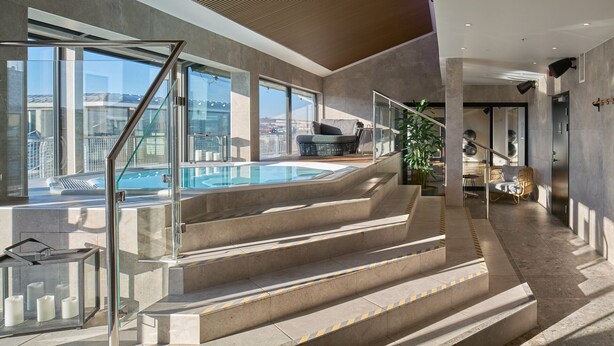 A modern indoor pool area with a glass railing, a staircase leading to a seating area, and a view of the cityscape. - Scandic Crown (Gothenburg)