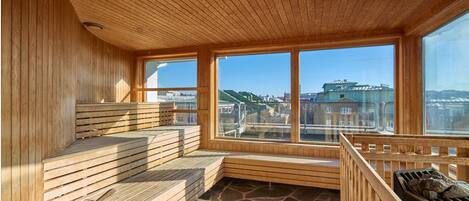 Una sala de sauna con bancos de madera, suelo de piedra y amplias ventanas con vistas a la ciudad.