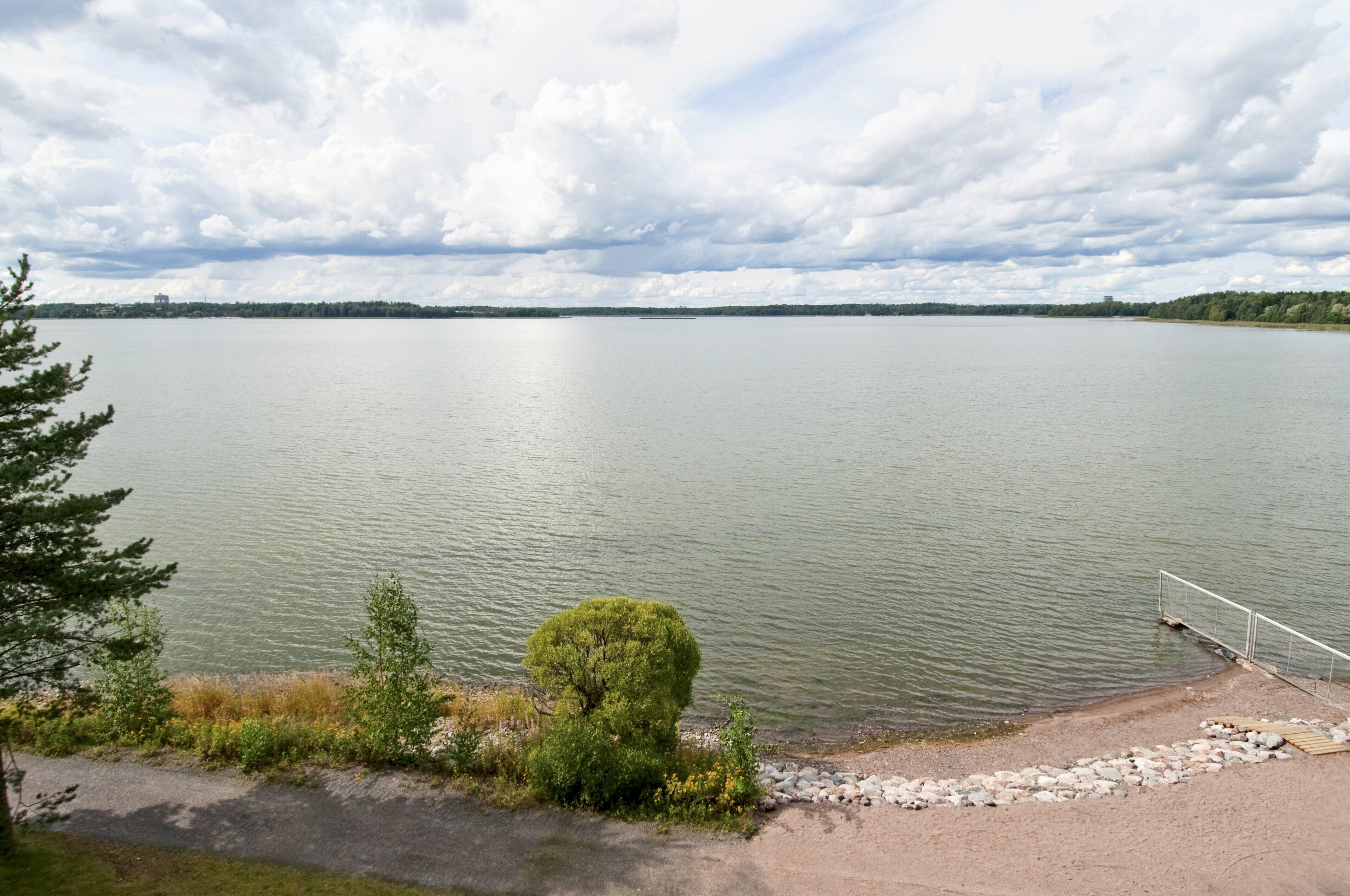 A calm lake with a sandy shore and a metal railing.