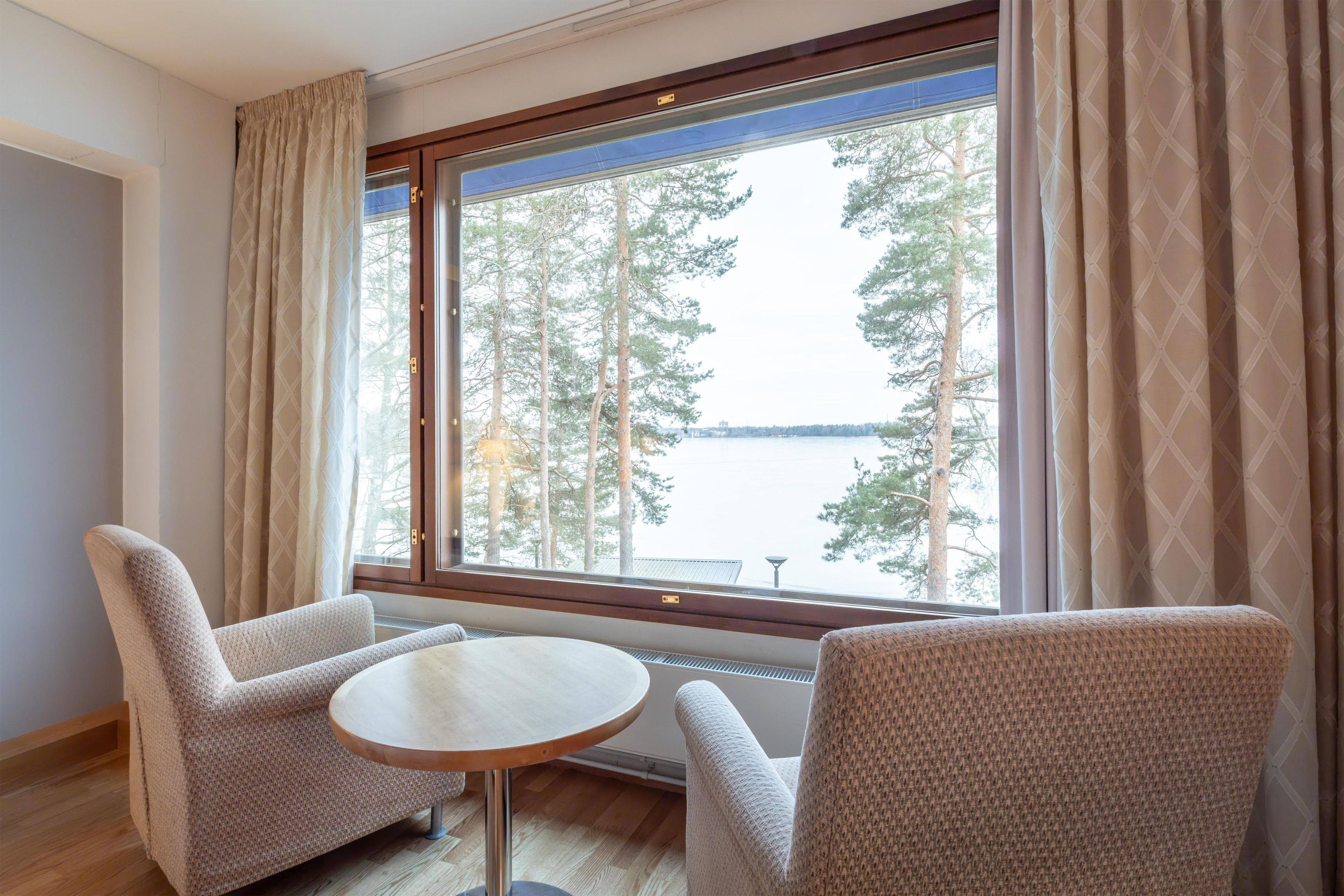 A room with two armchairs and a small round table, a large window with curtains, and a view of trees and a body of water.