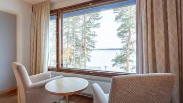 A room with two armchairs and a small round table, a large window with curtains, and a view of trees and a body of water.
