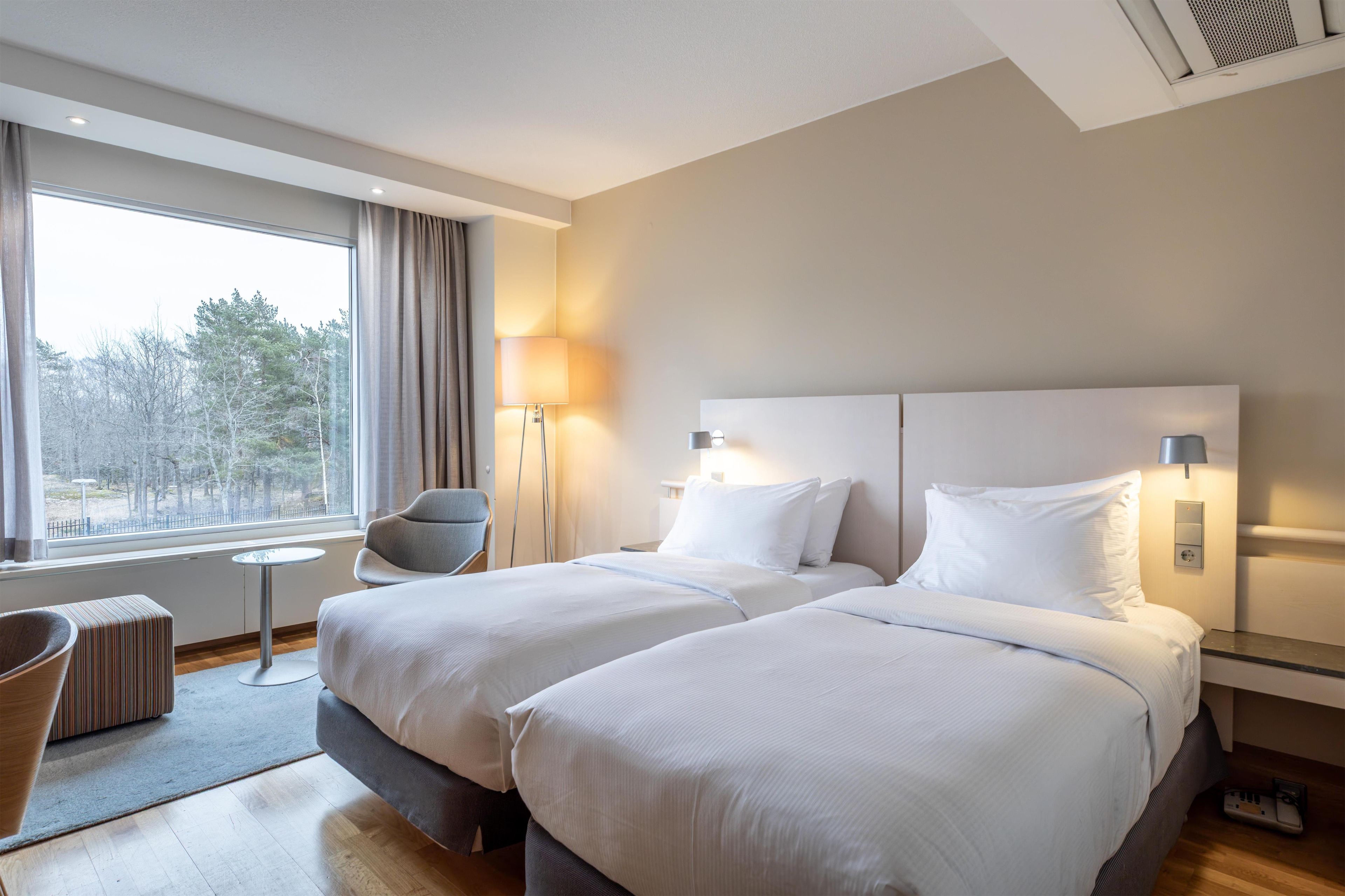 Quarto de hotel com duas camas, uma cadeira, uma mesinha e janela com vista para árvores.