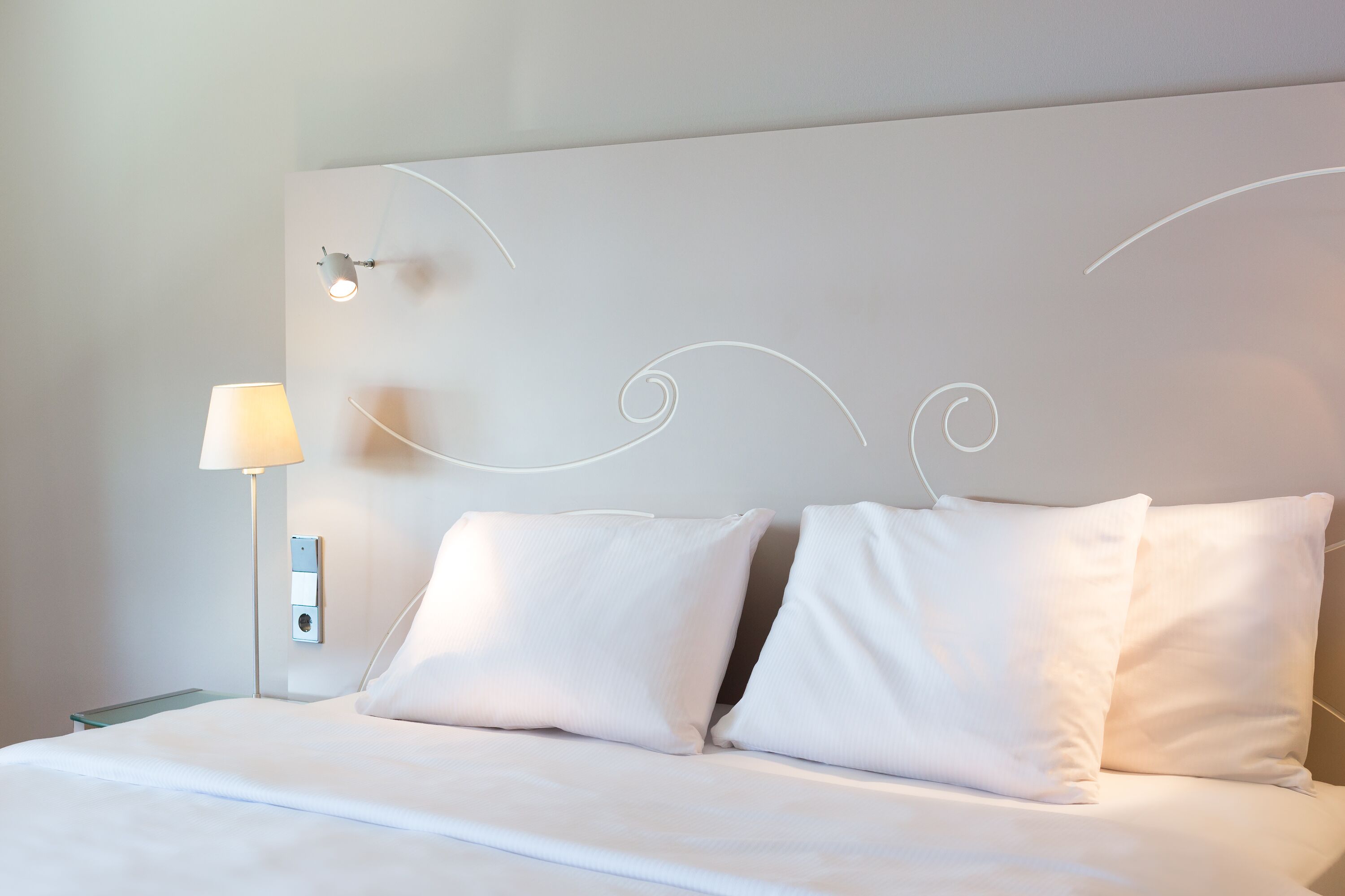 Un letto rifatto con lenzuola e cuscini bianchi, una lampada da comodino e un punto luce a parete.