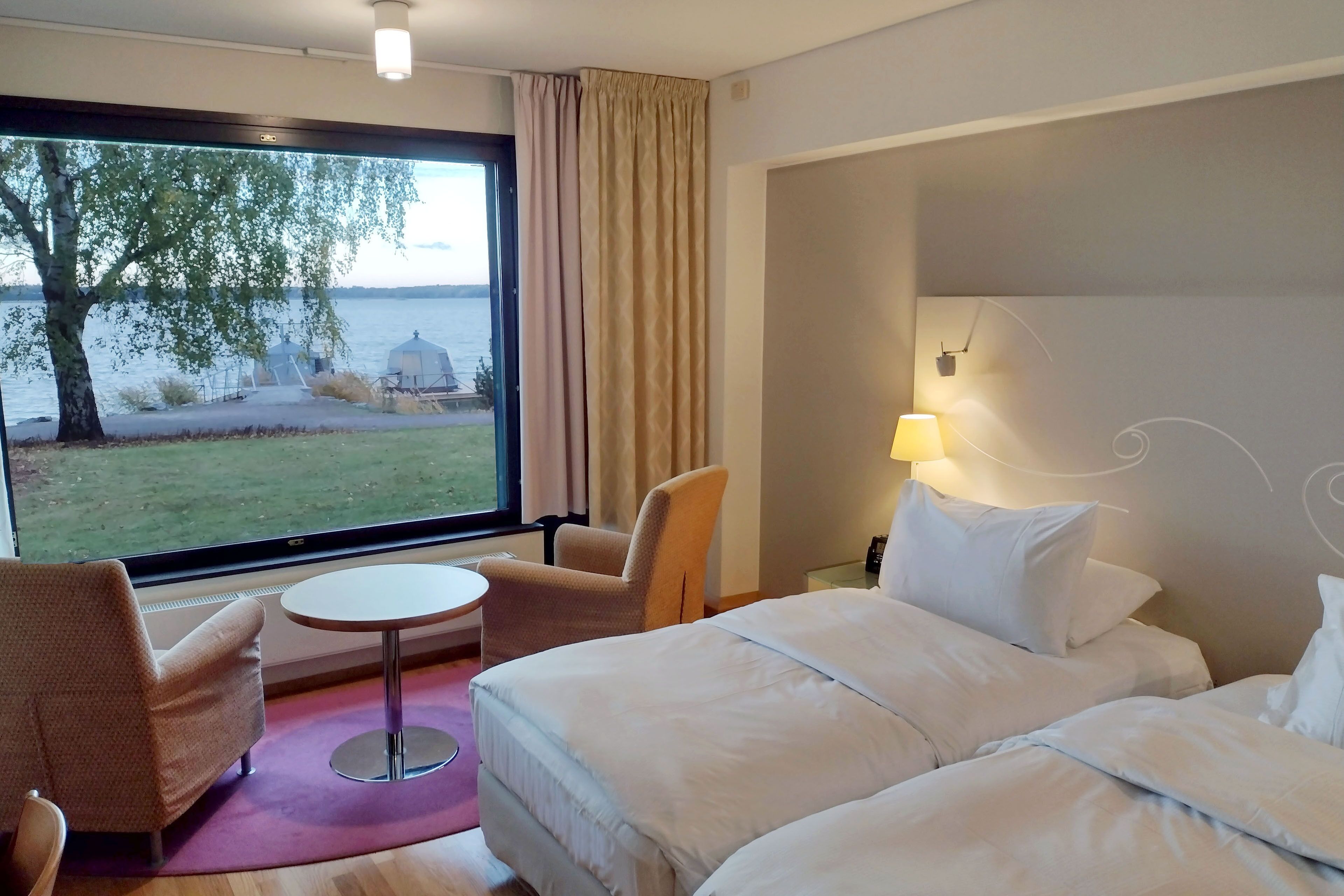 Ein Hotelzimmer mit großem Fenster, einem Bett, einem Sessel, einem kleinen Tisch und einer Lampe.