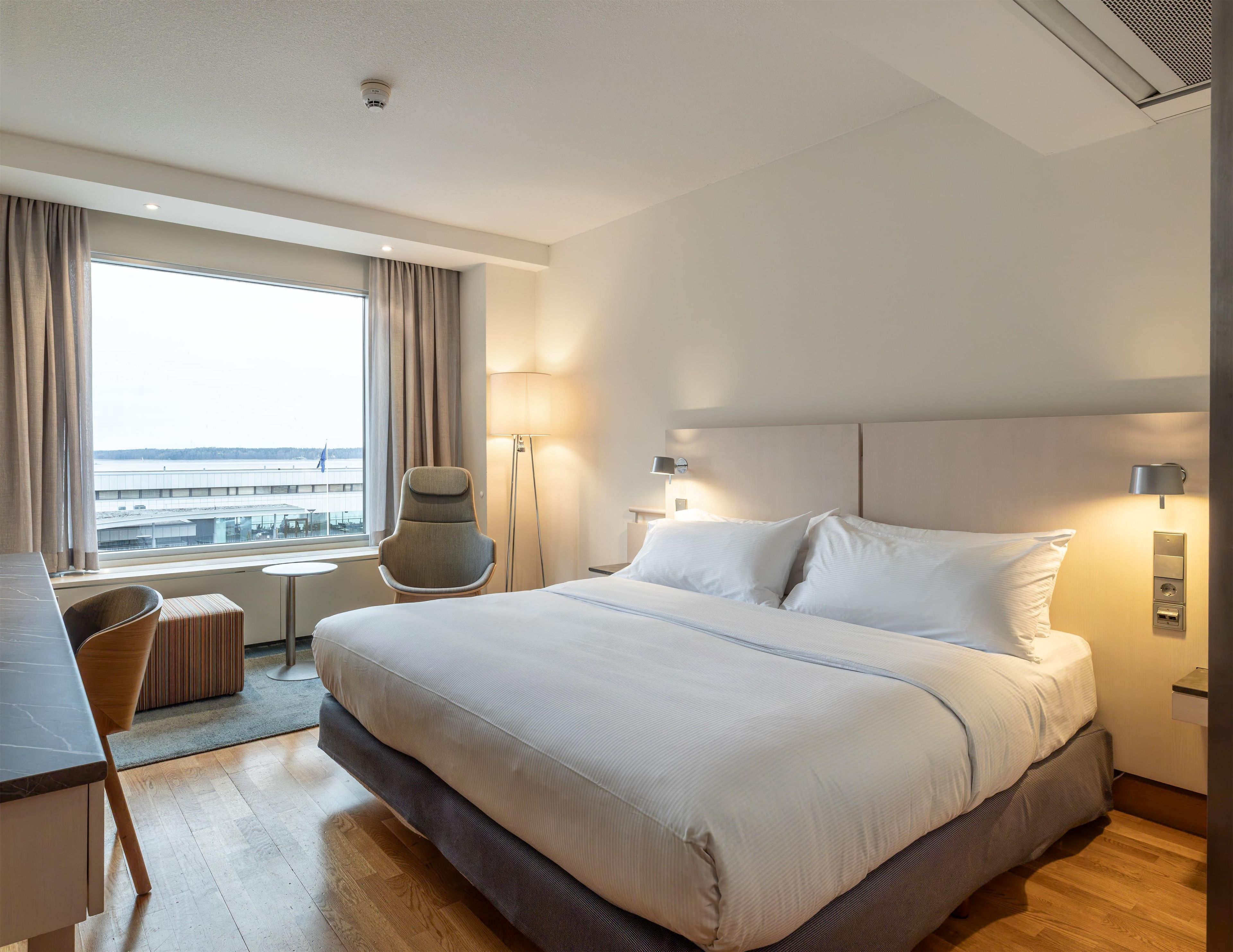 Une chambre d’hôtel avec un grand lit, un bureau, une chaise, une lampe sur pied et une vue sur le paysage urbain.