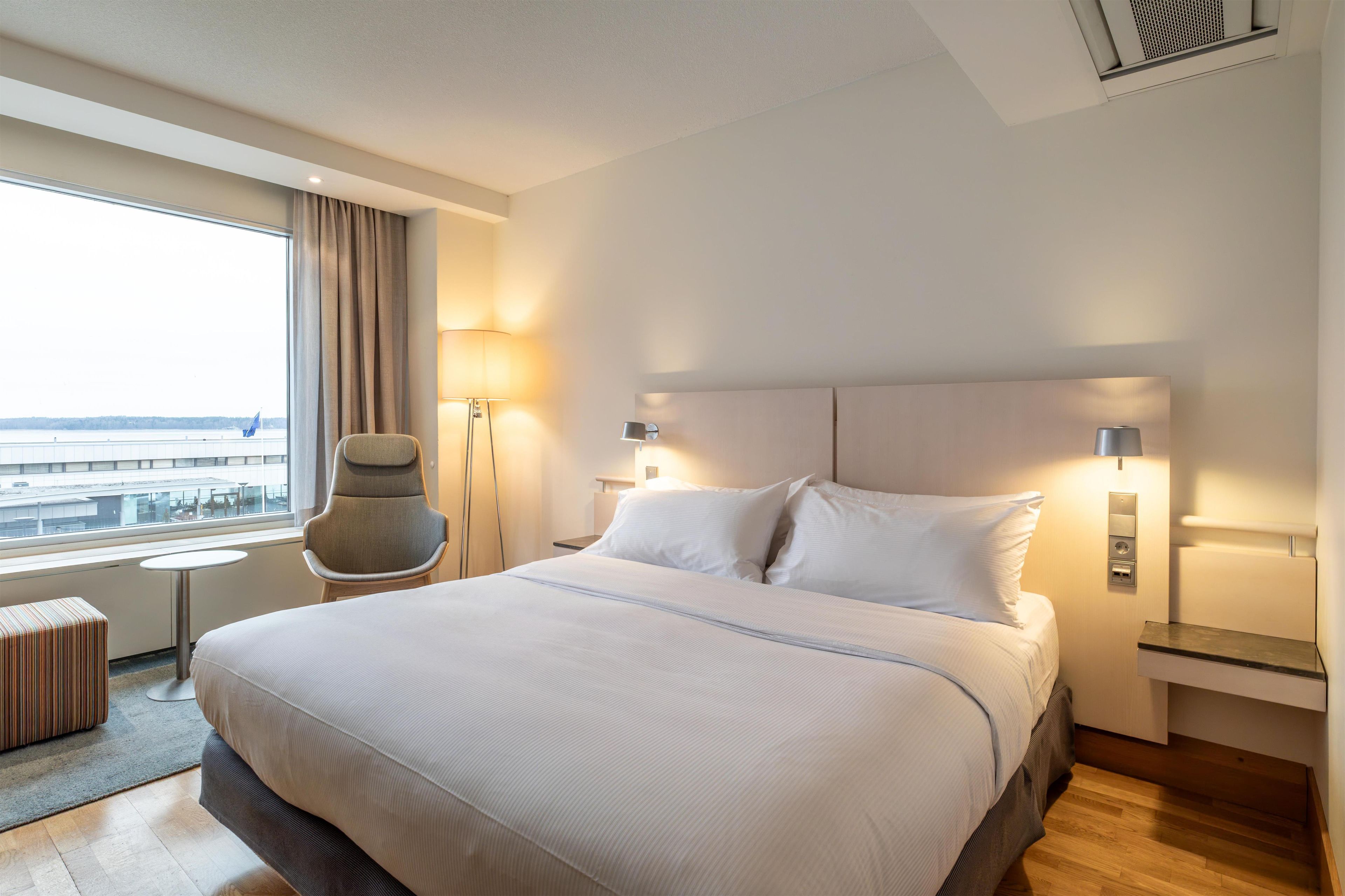 Habitación de hotel con una cama grande, una silla, una mesita y vista al aeropuerto.