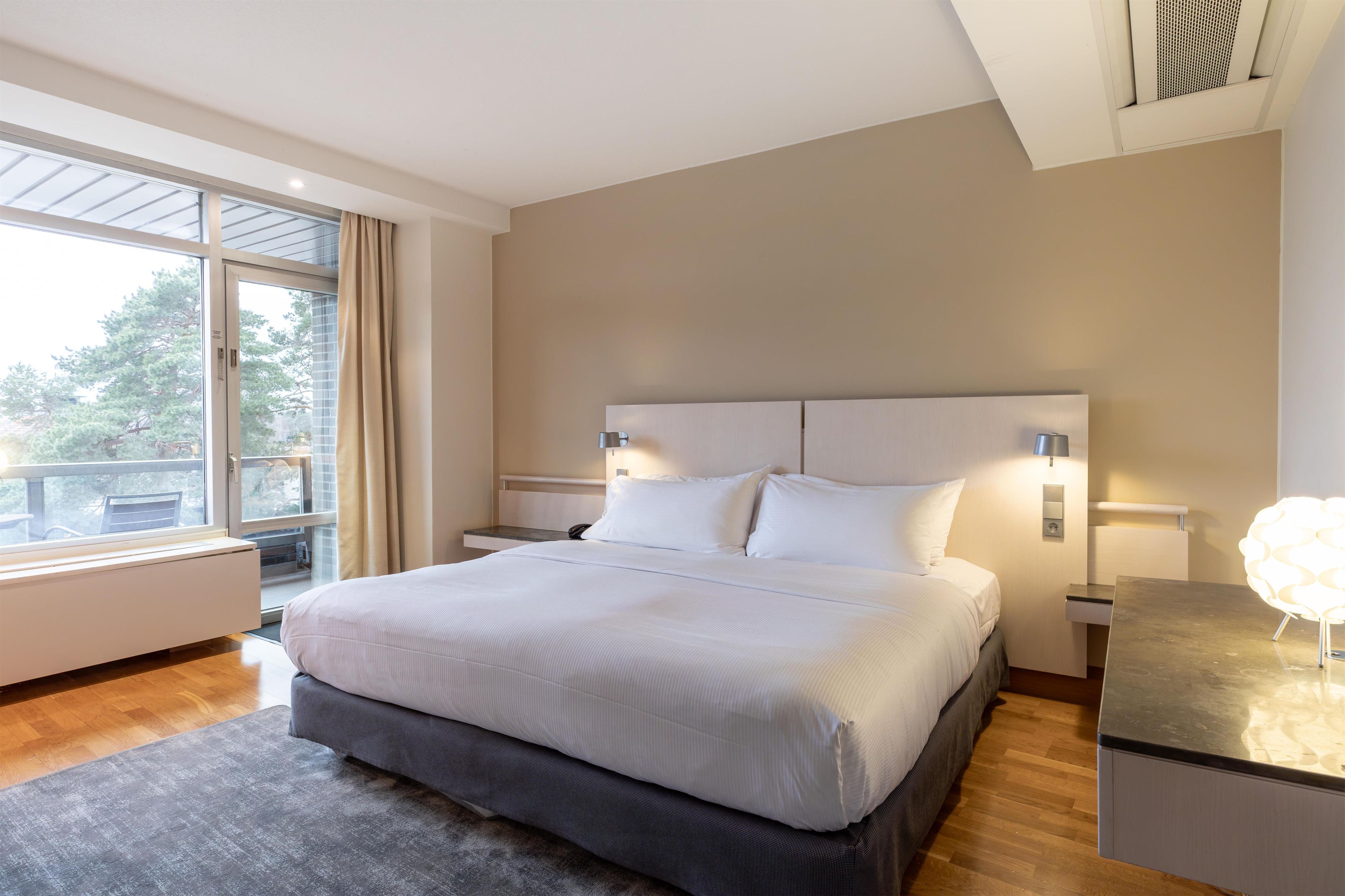 A modern hotel room with a large bed, a view of trees through the window, and a balcony.