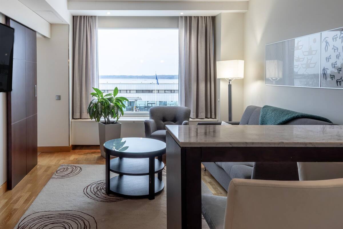 Ein modernes Hotelzimmer mit großem Fenster, einem Esstisch, einer Couch, einer Lampe und einem Blumentopf.