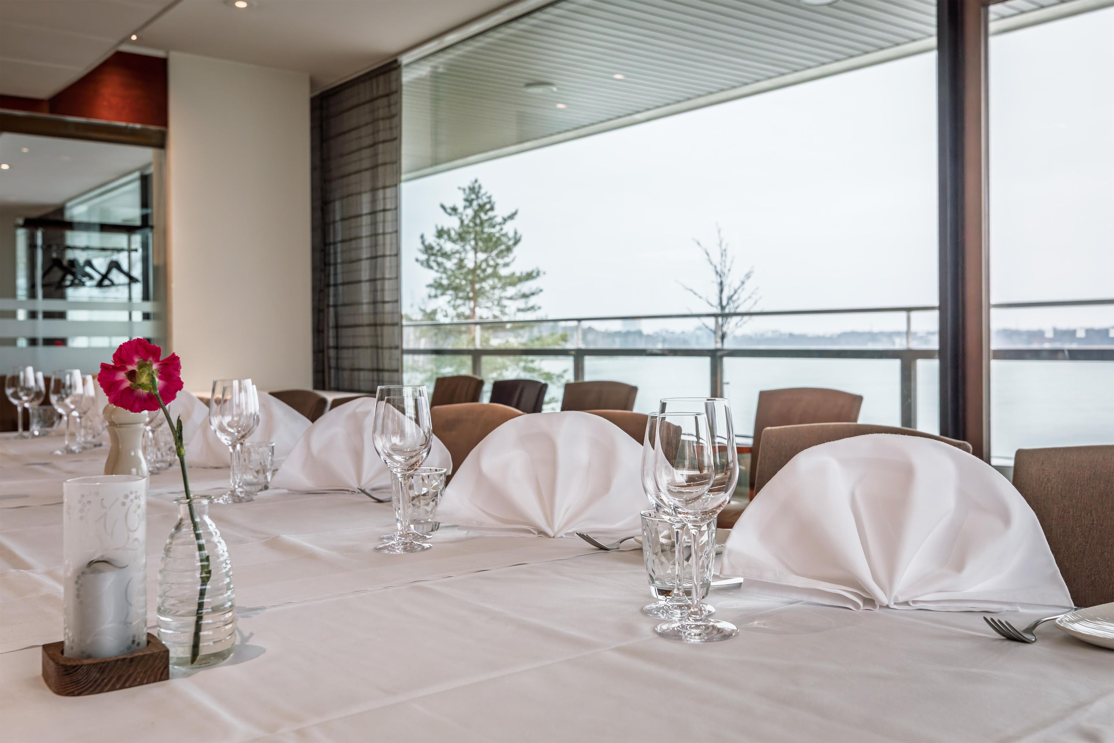 Une table à manger dressée avec des nappes blanches, de la verrerie et une seule fleur rose.