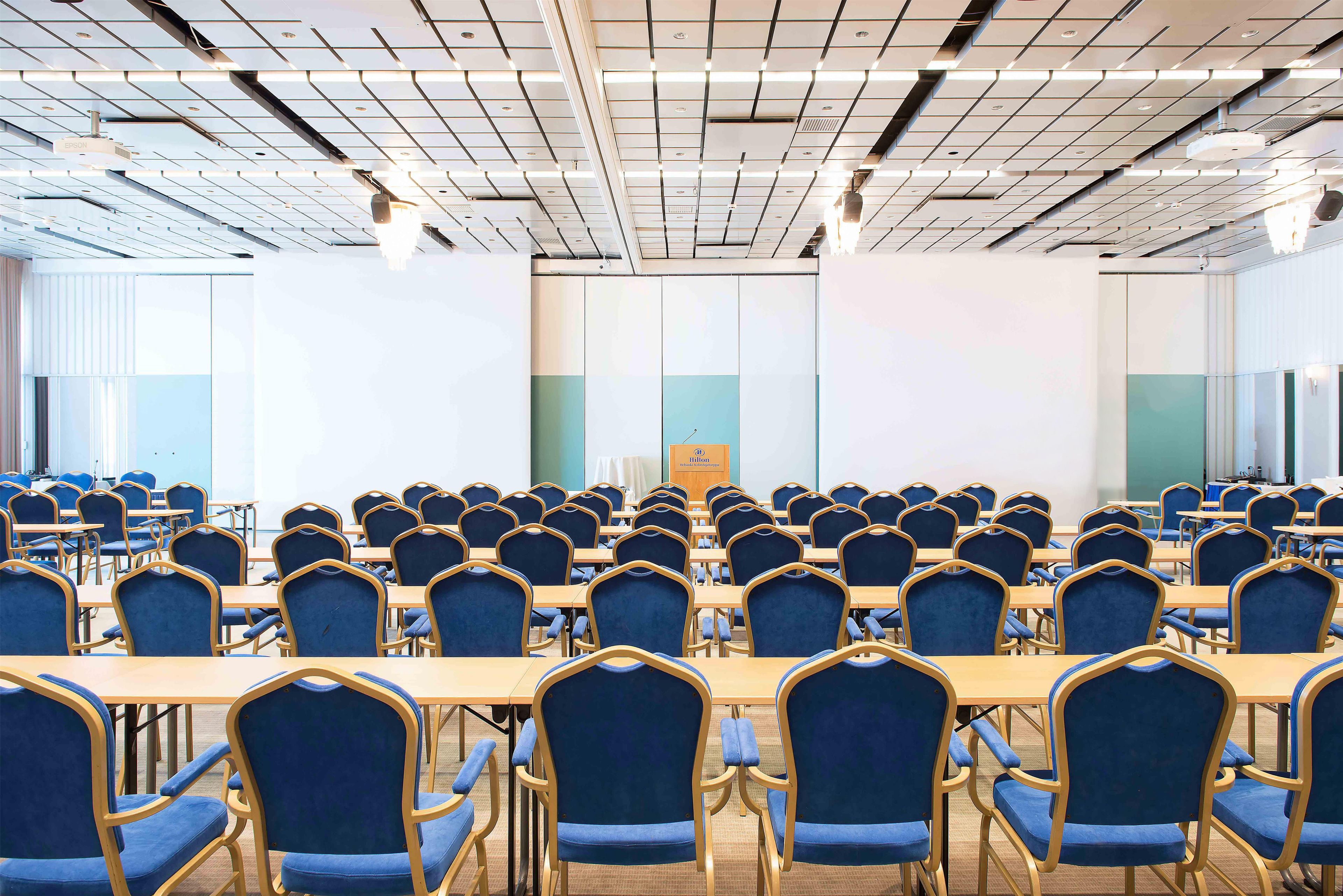 Un salón de conferencias con filas de mesas y sillas, un atril y un proyector en el techo.