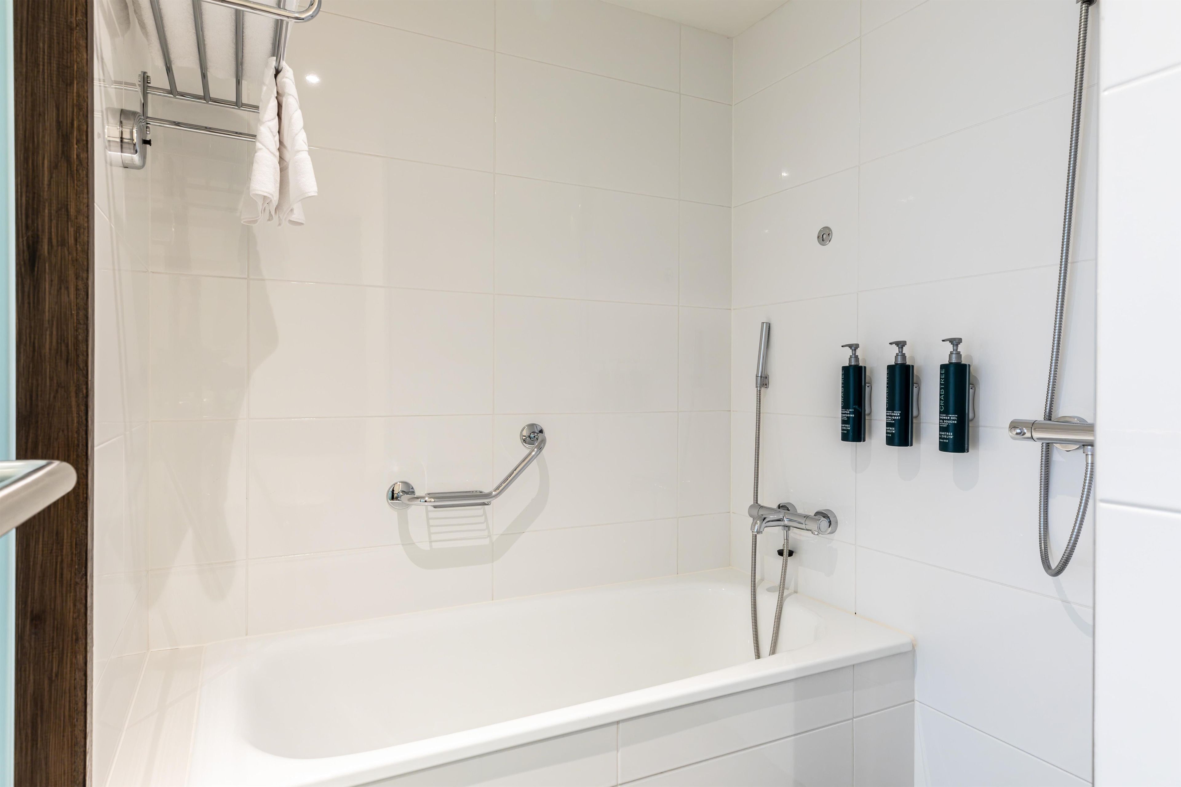 A modern bathroom with a white bathtub, a handheld showerhead, a grab bar, and three bottles of toiletries on the wall.