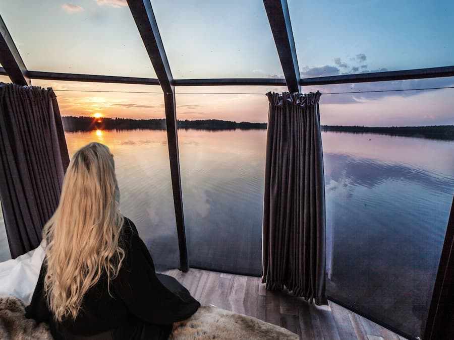 Una persona sentada junto a un ventanal grande con vistas a un lago tranquilo al atardecer.