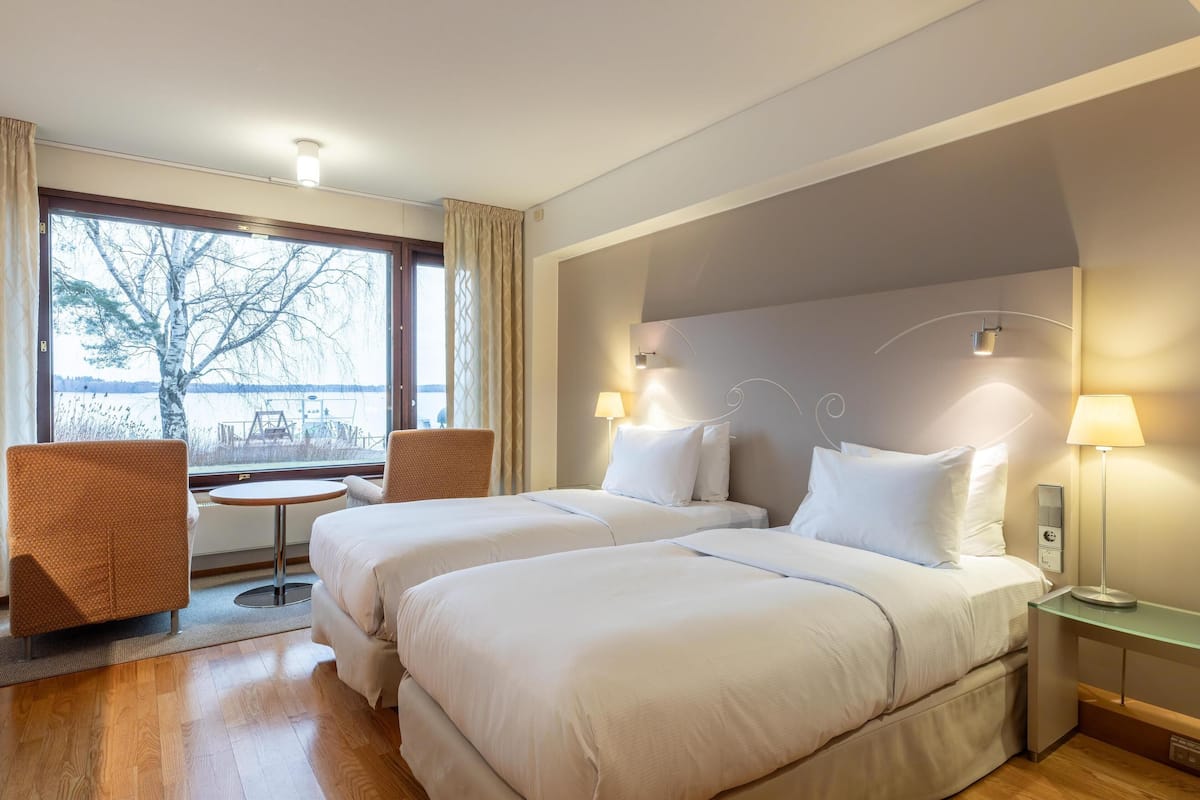 Ein Hotelzimmer mit zwei Betten, einem kleinen Tisch, einem Sessel und Blick auf einen Baum und einen See durch das Fenster.