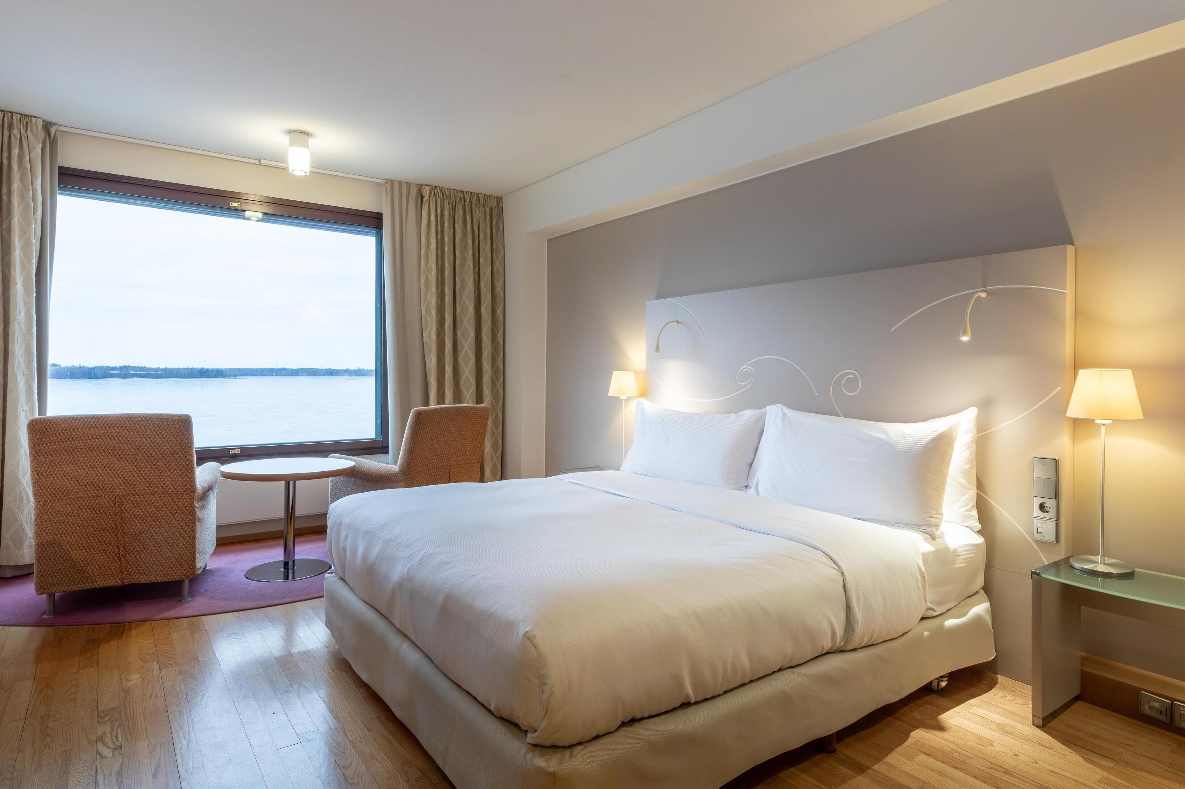 Une chambre d’hôtel avec un grand lit, une petite table ronde, deux chaises, un bureau avec une lampe et une vue sur l’eau.