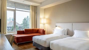Quarto de hotel com duas camas, um sofá, uma escrivaninha e uma janela grande com cortinas.