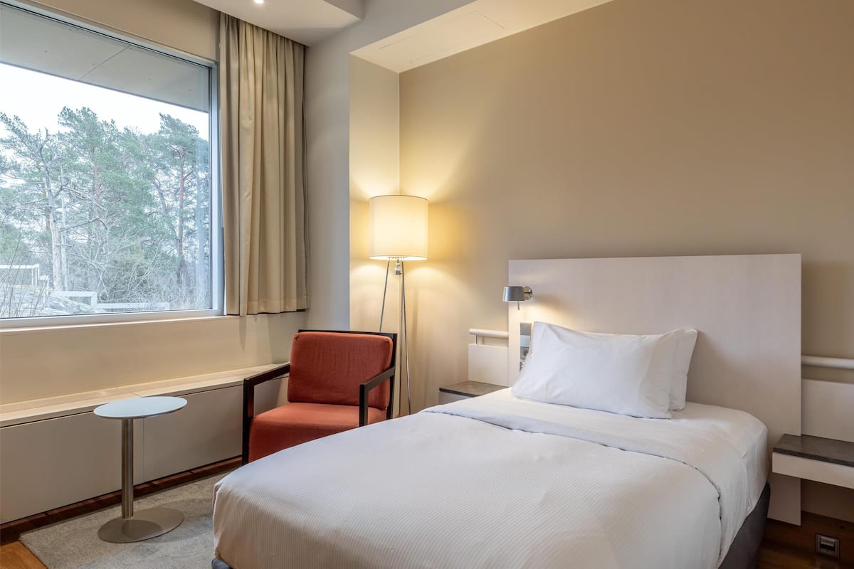 Ein Hotelzimmer mit einem großen Bett, einem Sessel, einem kleinen Tisch, einer Lampe und einem Fenster mit Vorhängen.
