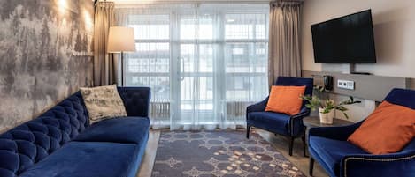 A modern living room with a blue sofa, two armchairs, a flat-screen TV, and a large window with curtains.