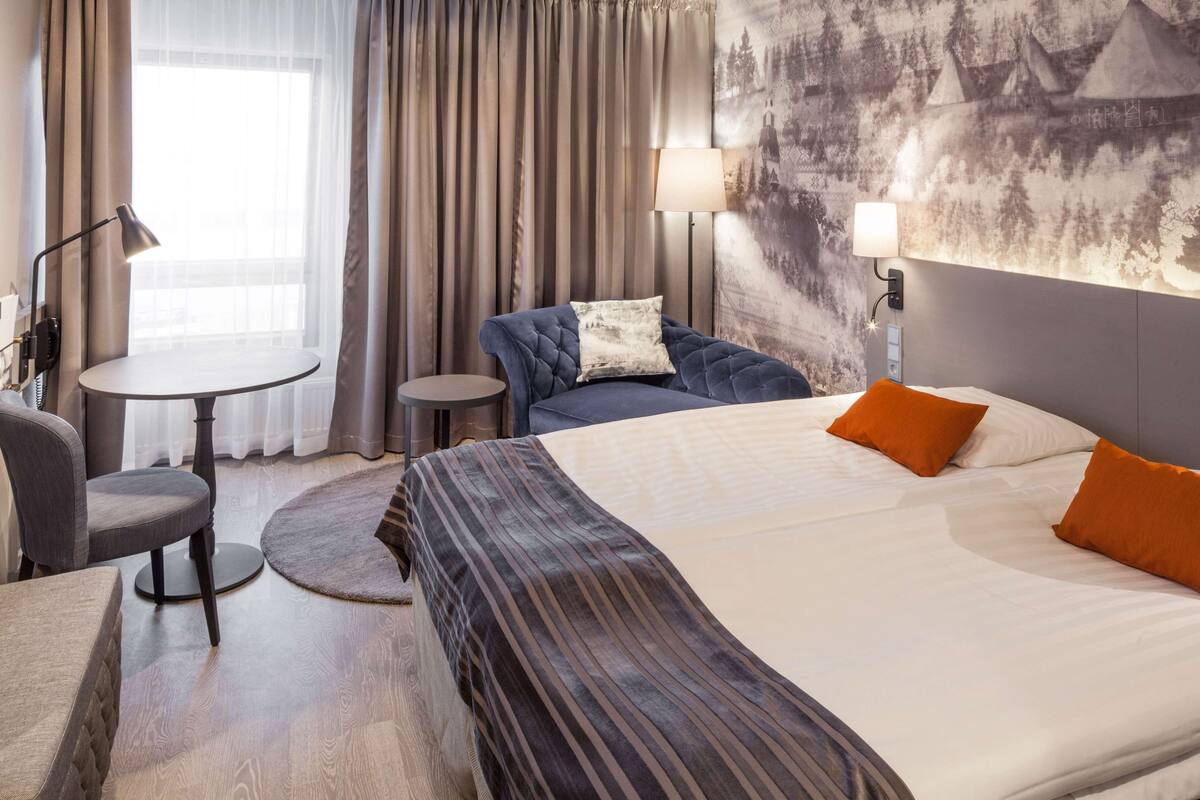 A hotel room with a large bed, a sofa, a small round table, and a chair. There is a window with curtains, a wall-mounted lamp, and a painting on the wall.
