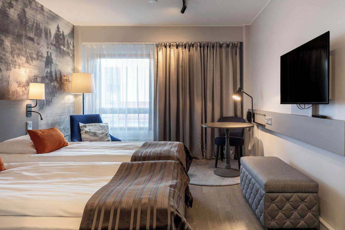 A hotel room with two beds, a desk, a chair, a TV, and a large window with curtains.