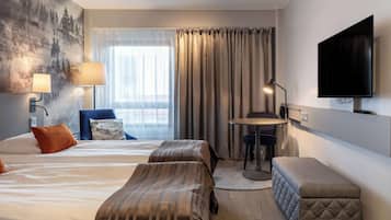 Ett hotellrum med två sängar, ett skrivbord, en stol, en TV och ett stort fönster med gardiner.