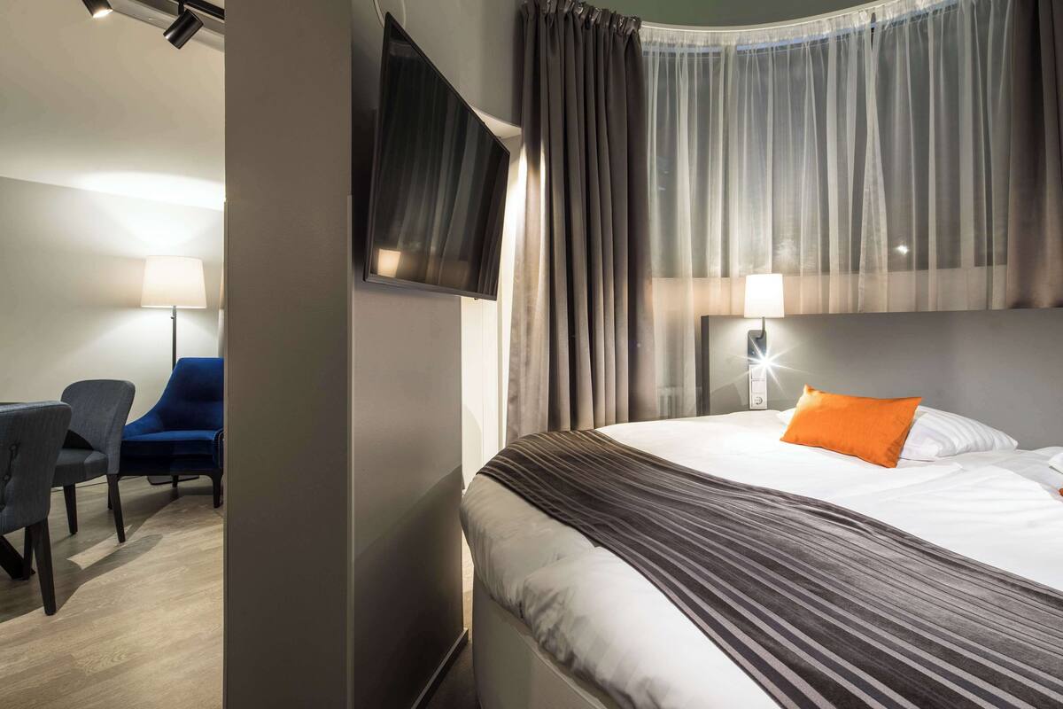 A hotel room with a bed, a desk with a chair, a TV mounted on the wall, and a lamp.
