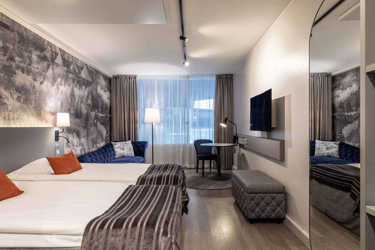 A hotel room with two beds, a desk, a chair, a small table, a mirror, and a window with curtains.