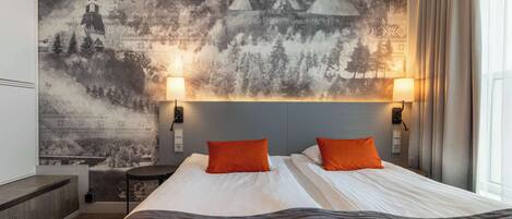 Habitación de hotel con una cama grande, dos almohadas naranjas, cabecero, lámparas de noche, un mural en la pared y una ventana con cortinas.