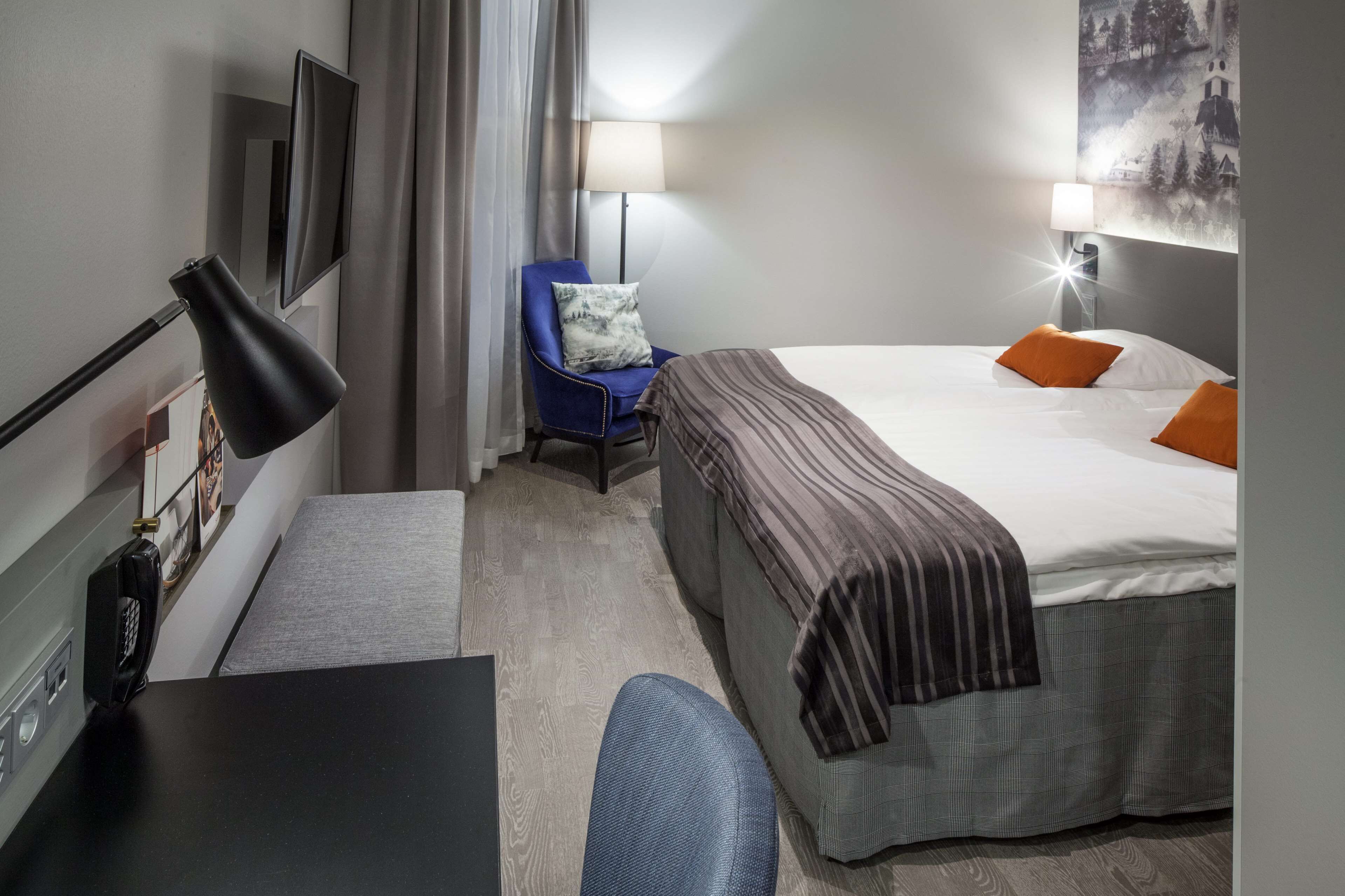 A hotel room with a bed, a desk, a chair, a lamp, and a painting on the wall.