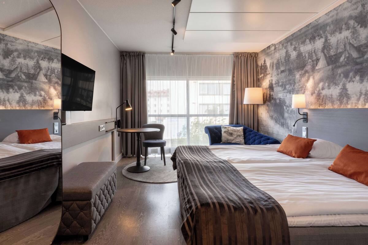 A hotel room with two beds, a desk, and a chair. There is a large window with curtains, a flat-screen TV mounted on the wall, and a lamp on the desk.